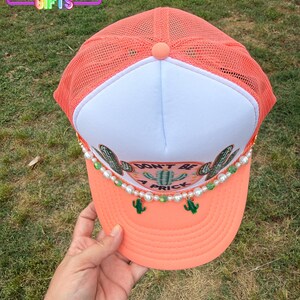 May include: Coral trucker hat with a white front panel and mesh back. The hat features a cactus design, the phrase "DON'T BE A PRICK", and pearl and green bead accents. The hat is from Rainbow Emporium Gifts.
