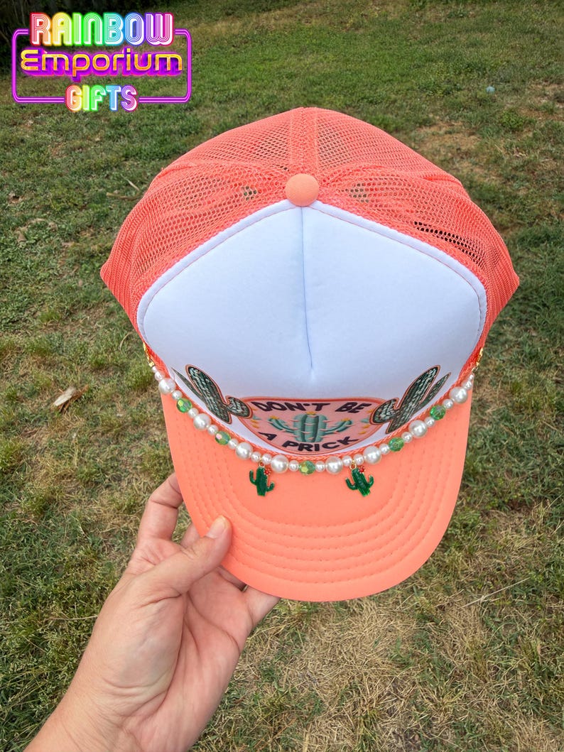 May include: Coral trucker hat with a white front panel and mesh back. The hat features a cactus design, the phrase "DON'T BE A PRICK", and pearl and green bead accents. The hat is from Rainbow Emporium Gifts.
