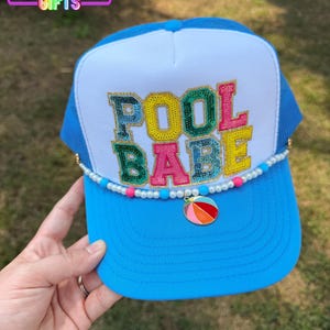 May include: A blue trucker hat with a white front panel. The hat features the text "POOL BABE" in colorful embroidered letters. A beaded strand with a beach ball charm adorns the brim. The top of the hat has the text "RAINBOW EMPORIUM GIFTS".