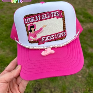 May include: Pink trucker hat with a white front panel featuring an embroidered image of a woman in a pink cowboy hat and a white shirt with the text "LOOK AT ALL THE FUCKS I GIVE". The hat has a white pearl trim and a pink cowboy hat charm.