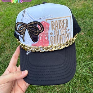 May include: Black trucker hat with a white top panel featuring a cowboy boot and bow design. The hat has the text "RAISED ON 90'S COUNTRY" and a gold chain across the brim. The Rainbow Emporium Gifts logo is visible.