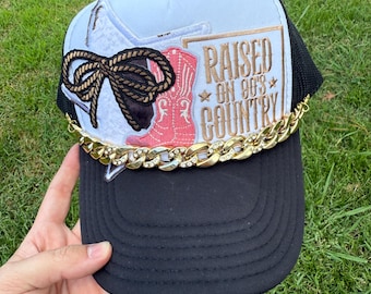 May include: Black trucker hat with a white top panel featuring a cowboy boot and bow design. The hat has the text "RAISED ON 90'S COUNTRY" and a gold chain across the brim. The Rainbow Emporium Gifts logo is visible.