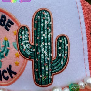 May include: A white trucker hat with a green sequined cactus patch. The hat has a coral mesh back and a beaded embellishment with pearls and green faceted beads. The text "BE" and "ICK" are visible on the hat.