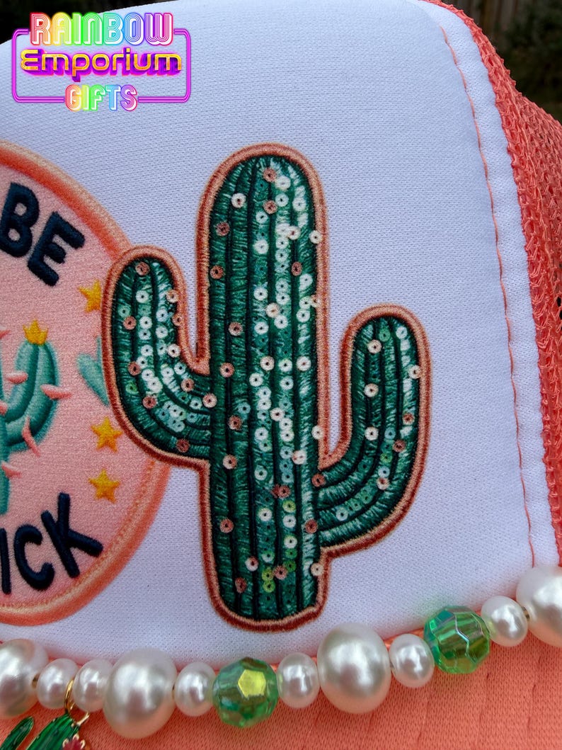 May include: A white trucker hat with a green sequined cactus patch. The hat has a coral mesh back and a beaded embellishment with pearls and green faceted beads. The text "BE" and "ICK" are visible on the hat.