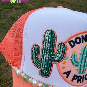 May include: A coral and white trucker hat with a sequined cactus patch and a beaded charm. The hat features a mesh back, a curved brim, and the text "DON'T BE A PRICK". The hat is from Rainbow Emporium Gifts.