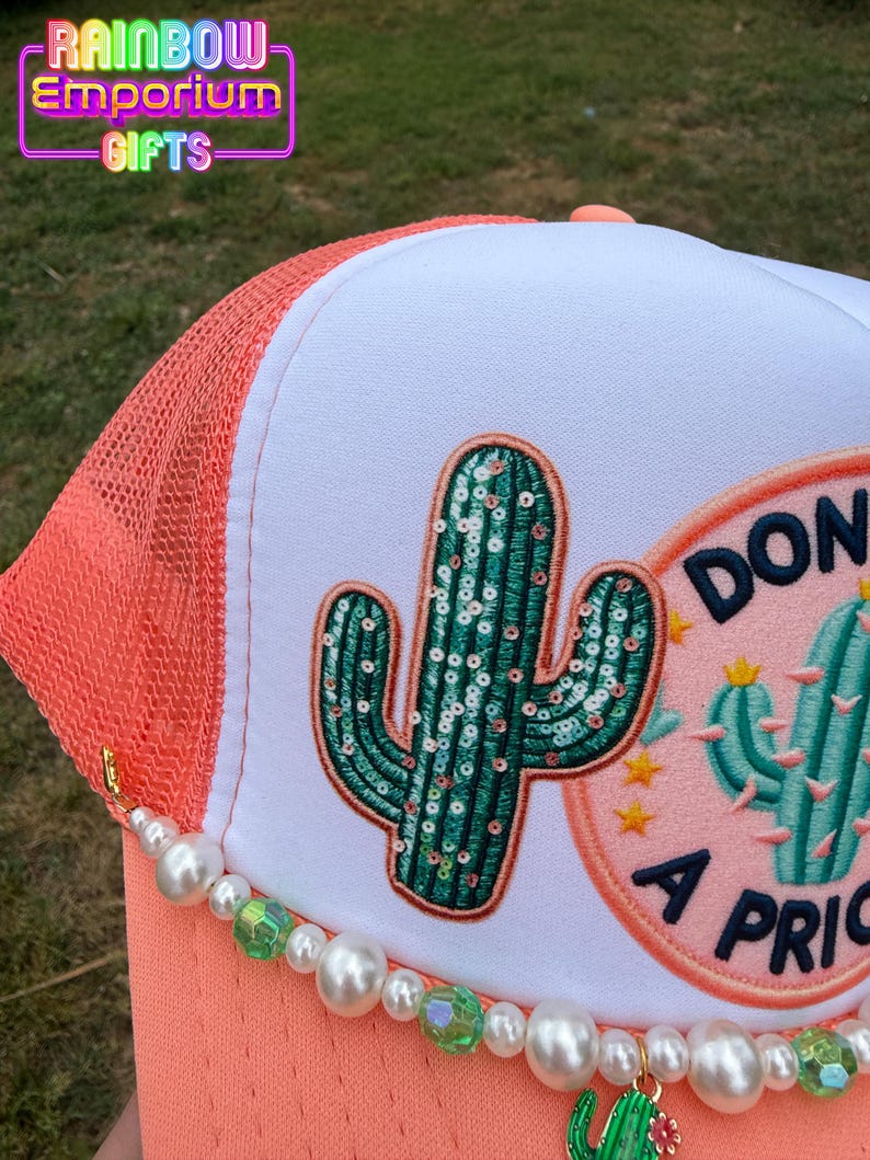 May include: A coral and white trucker hat with a sequined cactus patch and a beaded charm. The hat features a mesh back, a curved brim, and the text "DON'T BE A PRICK". The hat is from Rainbow Emporium Gifts.