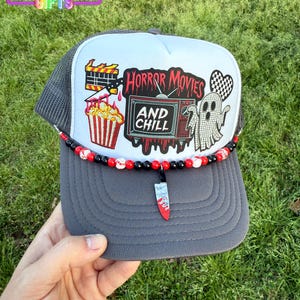 May include: A gray and white trucker hat with a horror movie theme. The hat features a ghost, a TV with the words "Horror Movies and Chill", popcorn, and a bloody knife. The hat has a black and white beaded trim.
