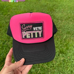 May include: A black and pink trucker hat with the text "Sorry We're Petty" on the front. The hat has a pink front panel, a black brim, and a black mesh back. The text is in a rectangular patch.