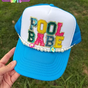 May include: A blue trucker hat with a white front panel. The hat features the text "POOL BABE" in colorful, beaded letters. A pearl and flower trim adorns the brim. The top of the hat has a blue mesh design. The text "RAINBOW EMPORIUM GIFTS" is at the top.