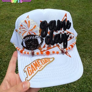 May include: White trucker hat with "Game Day" text and football-themed embellishments. Features a football, star, and beaded accents in orange, white, and black. The hat has a mesh back and a curved brim.