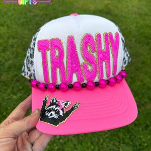 May include: A bright pink baseball cap with the word "TRASHY" in large, glittery pink letters. The cap features a black and white leopard print mesh, a pink brim, and a raccoon patch. The text "Rainbow Emporium Gifts" is at the top.