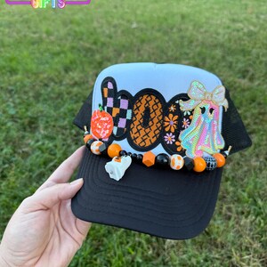 May include: Black and white Halloween-themed trucker hat with the word "BOO" in orange, purple, and black. The hat features a ghost, pumpkin, and beaded accents. The brim is black, and the crown is white with black mesh.