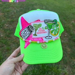 May include: A neon green trucker hat with a white front panel and a green mesh back. The hat is decorated with a pink star, a margarita glass, a lime slice, and the text "It's 5 O'Clock Somewhere".