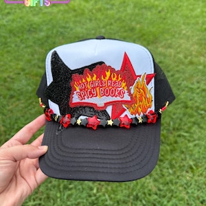 May include: Black trucker hat with a white panel featuring a star, flames, and the text "HOT GIRLS READ SPICY BOOKS." The brim is black, and the hat is decorated with red star-shaped beads.