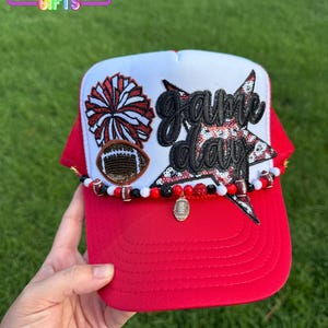 May include: Red and white trucker hat with a football theme. The hat features a red brim and a white front panel with the words "Game Day" in black script. Embellishments include a football, a pom-pom, and a beaded trim.