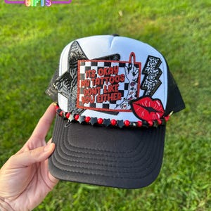 May include: Black and white trucker hat with a mesh back and a curved brim. The hat features the text "IT'S OKAY MY TATTOOS DON'T LIKE YOU EITHER" with a hand gesture, a lightning bolt, and red lips. The hat is decorated with black and red beads.