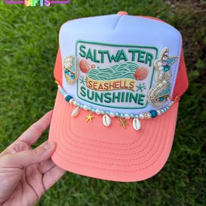 May include: A coral and white trucker hat featuring a central embroidered patch with the text SALTWATER SEASHELLS SUNSHIINE, surrounded by beaded seashell and starfish embellishments, and a dangling trim of pearls, blue beads, and charms.