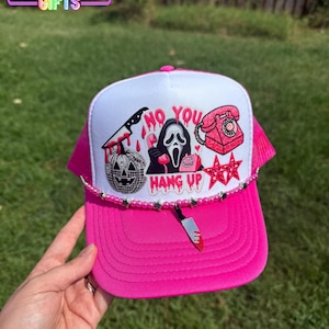 May include: A bright pink and white trucker hat featuring a graphic print of a ghost mask, a disco ball pumpkin, a pink rotary phone, and a knife with the text NO YOU HANG UP. The hat is adorned with a pink beaded trim and a small knife charm.