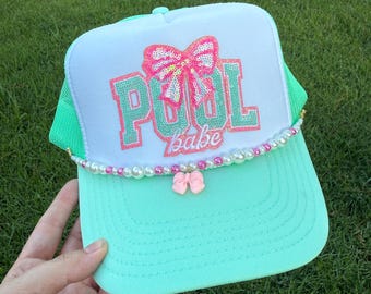 May include: A mint green and white trucker hat with the words "POOL babe" in sequined letters and a pink sequined bow. The hat has a pearl and pink bow accent on the brim. The top of the hat has the text "RAINBOW Emporium GIFTS".