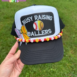 May include: Black and white baseball cap with a patch that reads "BUSY RAISING BALLERS" and a baseball graphic. The cap has a beaded trim with baseball and red and yellow beads. The cap is held up by a hand.