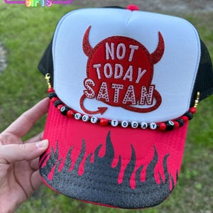 May include: A red and black trucker hat with a white mesh back. The front of the hat features a red devil with horns and the text "Not Today Satan". The brim of the hat has a black flame design.