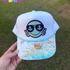 May include: White trucker hat with a mesh back and a curved brim. The front features a smiling face with sunglasses and peace signs. The brim has a beach scene with seashells and a beaded accent. The text "RAINBOW Emporium GIFTS" is visible.