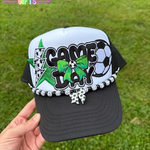 May include: Black and white trucker hat with "GAME DAY" text, soccer ball and star graphics. The hat has a black brim, white front panel, and black mesh back. A decorative beaded trim and a bow accent the brim.