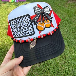 May include: A white and red trucker hat with a black brim featuring a black and white checkered patch with the text Game Day. An embroidered black and white bow sits above two orange footballs. A beaded strand with football charms lines the brim.