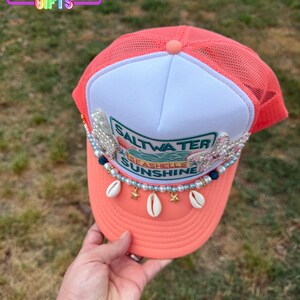 May include: Coral-colored trucker hat with a white front panel. The hat features the text "Saltwater, Seashells, Sunshine" and is adorned with seashells, pearls, and star charms. The top of the hat has a mesh back.