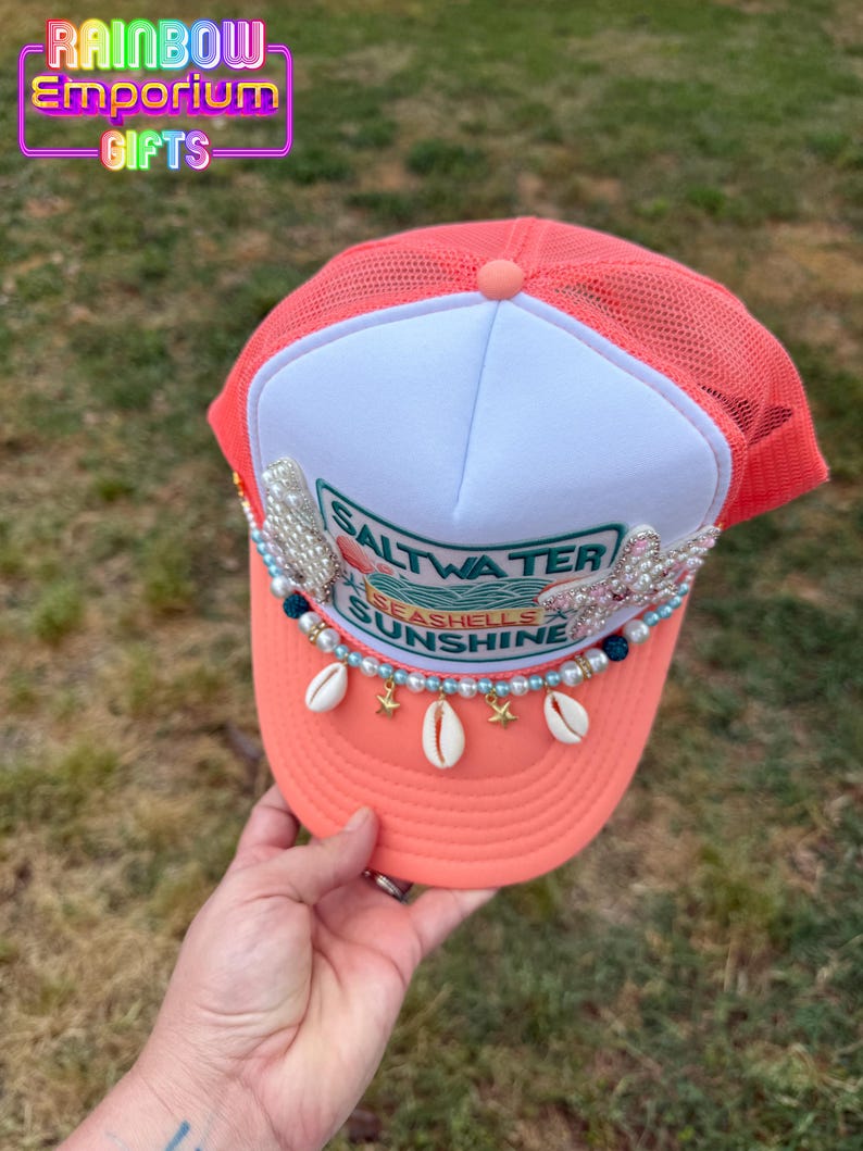 May include: Coral-colored trucker hat with a white front panel. The hat features the text "Saltwater, Seashells, Sunshine" and is adorned with seashells, pearls, and star charms. The top of the hat has a mesh back.