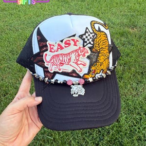 May include: A black trucker hat with a white and black mesh back. The hat features a red and white tiger patch with the word "EASY", a gold and black tiger patch, and a star-shaped patch. A silver tiger charm hangs from the brim.