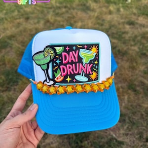 May include: A blue and white trucker hat with a colorful embroidered patch that says "Day Drunk" and features a margarita glass. The hat has a row of orange star-shaped beads around the brim.