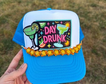 May include: A blue and white trucker hat with a colorful embroidered patch that says "Day Drunk" and features a margarita glass. The hat has a row of orange star-shaped beads around the brim.