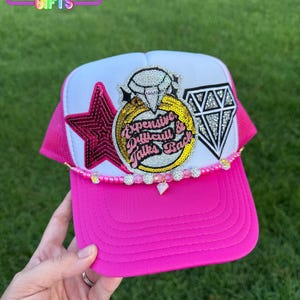 May include: A hot pink trucker hat with a white front panel and mesh back. The hat features a star, diamond, and ring design with the text "Expensive, Difficult & Takes Back". Embellished with rhinestones.