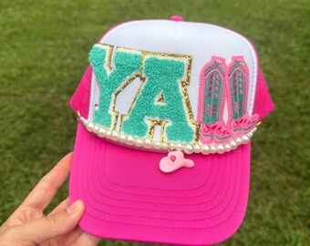 May include: A pink and white trucker hat with the word "Y'ALL" in green and gold. The hat features a pink brim, a white top, and a pink mesh back. It is decorated with a pearl trim and a small pink cowboy hat charm.