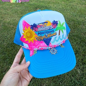 May include: A blue trucker hat with a white front panel featuring a colorful embroidered design of a sunset, palm trees, and the words "Pontoon & High Noons." The hat has a curved brim and a beaded embellishment with charms.