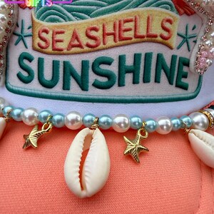 May include: A coral-colored hat with the words "SEASHELLS SUNSHINE" embroidered on the front. The hat is adorned with pearls, seashells, and gold starfish charms. The top of the hat has a white and green design.