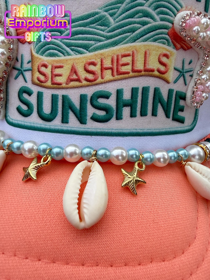 May include: A coral-colored hat with the words "SEASHELLS SUNSHINE" embroidered on the front. The hat is adorned with pearls, seashells, and gold starfish charms. The top of the hat has a white and green design.
