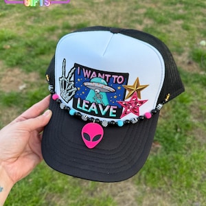 May include: Black and white trucker hat with a white patch that says "I WANT TO LEAVE" with a skeleton and a UFO. The patch has a pink, blue, and green color scheme. The hat has a black mesh back and a pink alien charm.