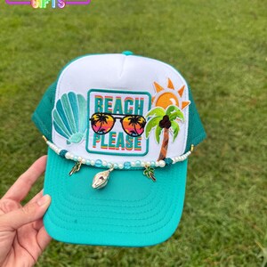 May include: A teal and white trucker hat with a beach theme. The hat features a seashell, sunglasses, palm tree, and sun design. The text "BEACH PLEASE" is displayed on the front. The hat is decorated with beads and charms. The logo "RAINBOW EMPORIUM GIFTS" is in the top left corner.