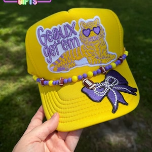 May include: A bright yellow trucker hat featuring an embroidered patch of a tiger wearing heart-shaped sunglasses with the text Geaux get 'em. The hat is decorated with a beaded string of yellow and purple beads, football charms, and a purple bow.