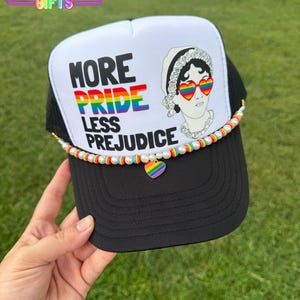 May include: Black and white trucker hat with the text "MORE PRIDE LESS PREJUDICE" and a graphic of a woman wearing rainbow heart-shaped sunglasses. The hat is decorated with a rainbow and pearl beaded chain and a rainbow heart charm.