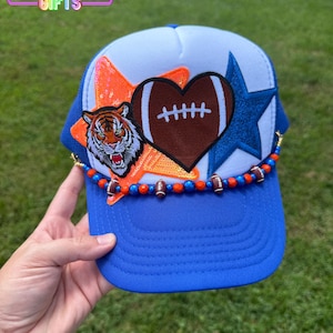 May include: A blue and white trucker hat with a football theme. The hat features a tiger patch, a football-shaped heart, and a blue star. The brim is blue, and the hat is decorated with orange and blue beads and football charms. The text "RAINBOW Emporium GIFTS" is visible.
