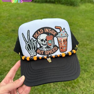 May include: Black and white trucker hat with the text "DEAD INSIDE BUT SPICED" and a skull graphic. The hat features a beaded accent with orange and black beads. The hat is being held up in front of a green grassy background.
