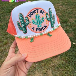 May include: A coral and white trucker hat with a mesh back. The front features a patch with a cactus design and the words "DON'T BE A PRICK". The brim is coral, and the hat is decorated with pearls and cactus charms. The text "RAINBOW EMPORIUM GIFTS" is at the top.
