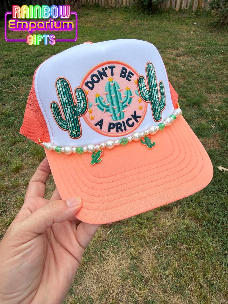 May include: A coral and white trucker hat with a mesh back. The front features a patch with a cactus design and the words "DON'T BE A PRICK". The brim is coral, and the hat is decorated with pearls and cactus charms. The text "RAINBOW EMPORIUM GIFTS" is at the top.