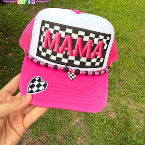 May include: Pink trucker hat with a black and white checkered patch that says "MAMA" in pink letters. The hat has a black and white checkered heart charm and black and white beads around the brim.