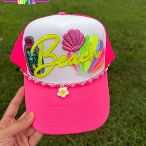 May include: A bright pink trucker hat with a white front panel. The hat features the word "Beach" in neon yellow script, a pink seashell, and colorful beach-themed graphics. A beaded chain with a white flower charm adorns the brim.