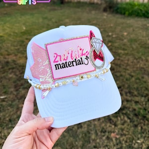 May include: White trucker hat with a mesh back and a curved brim. The front features a pink rectangle with the text "2nd Wife material" and a diamond ring design. Embellished with pearls, stars, and a pink wing.