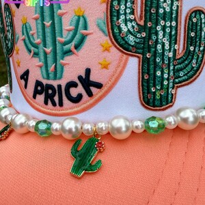 May include: A white and peach-colored hat with a beaded necklace. The hat features a cactus design and the text "A Prick". The necklace has a green cactus charm and pearl beads.